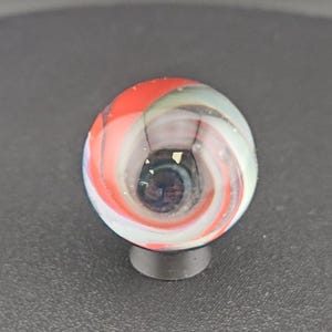 Könnte beinhalten: Eine kugelförmige Glasmurmel mit einem wirbelnden Muster aus undurchsichtigen roten, weißen und durchscheinenden blauen Bändern. Die Mitte der Murmel enthält einen dunklen, konzentrierten Kern mit kleinen, im Glas sichtbaren goldfarbenen Partikeln.
