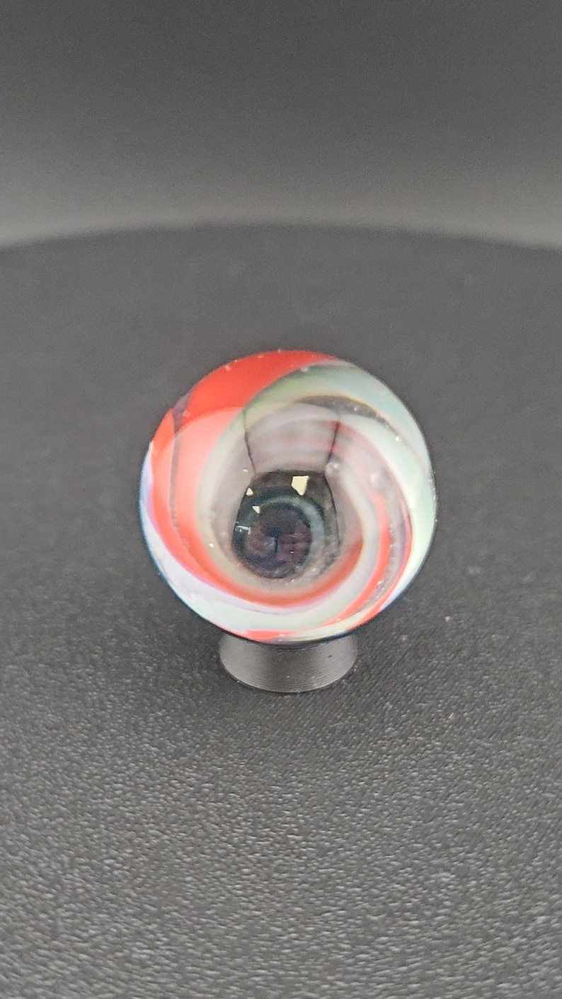 May include: A spherical glass marble featuring a swirling pattern of opaque red, white, and translucent blue ribbons. The center of the marble contains a dark, concentrated core with small flecks of gold-colored material visible within the glass.