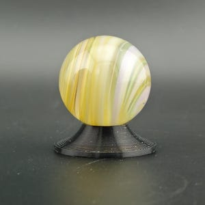 May include: A decorative glass marble with yellow, white, and green swirls, resting on a black, spiral-shaped base. The marble is spherical and appears translucent, with light passing through the colorful patterns.