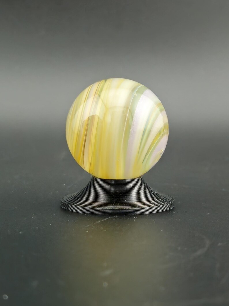May include: A decorative glass marble with yellow, white, and green swirls, resting on a black, spiral-shaped base. The marble is spherical and appears translucent, with light passing through the colorful patterns.