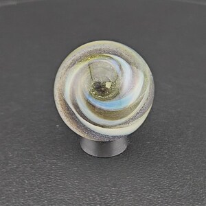 Puede incluir: Una canica de cristal esf&eacute;rica con un patr&oacute;n de remolinos en tonos blancos, dorados y azules iridiscentes. El interior contiene finas part&iacute;culas de purpurina met&aacute;lica que captan la luz, creando un efecto complejo y estratificado.