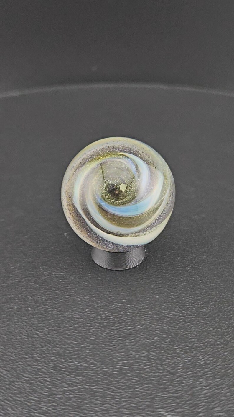 Puede incluir: Una canica de cristal esf&eacute;rica con un patr&oacute;n de remolinos en tonos blancos, dorados y azules iridiscentes. El interior contiene finas part&iacute;culas de purpurina met&aacute;lica que captan la luz, creando un efecto complejo y estratificado.