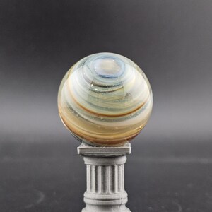 May include: A decorative glass orb with swirling patterns of blue, gold, and brown, resting on a small, gray, classical-style pedestal. The orb is spherical and appears to be made of glass.