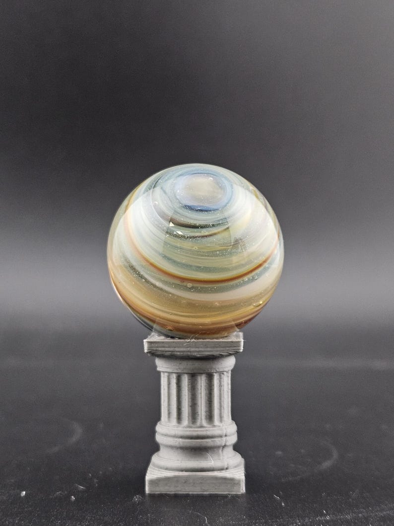 May include: A decorative glass orb with swirling patterns of blue, gold, and brown, resting on a small, gray, classical-style pedestal. The orb is spherical and appears to be made of glass.
