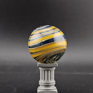 May include: A decorative glass sphere with swirling patterns of yellow, blue, and brown, resting on a small, gray, fluted pedestal. The sphere has a glossy finish and appears to be made of glass. The pedestal is square and has a classical design.