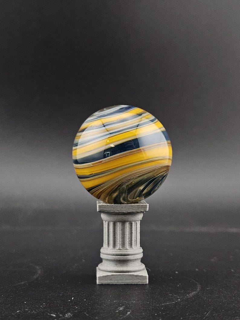 May include: A decorative glass sphere with swirling patterns of yellow, blue, and brown, resting on a small, gray, fluted pedestal. The sphere has a glossy finish and appears to be made of glass. The pedestal is square and has a classical design.