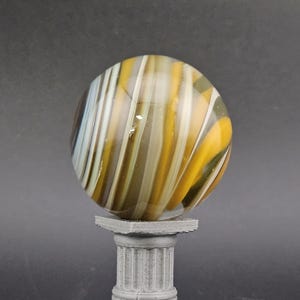 May include: A decorative glass marble with yellow, white, and brown swirling stripes, resting on a small, gray, fluted column pedestal. The marble is spherical and appears translucent.