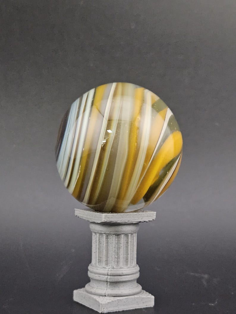 May include: A decorative glass marble with yellow, white, and brown swirling stripes, resting on a small, gray, fluted column pedestal. The marble is spherical and appears translucent.