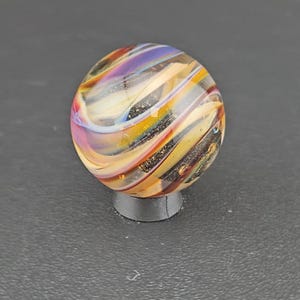 May include: A spherical glass marble featuring swirling patterns of purple, yellow, orange, and white colors. The surface of the marble contains embedded dark glitter particles that catch the light, creating a multi-colored effect throughout the glass.