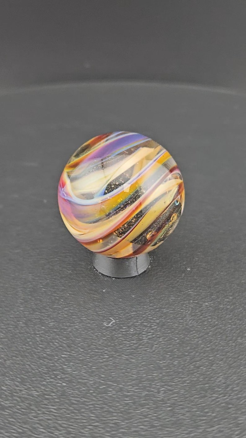 May include: A spherical glass marble featuring swirling patterns of purple, yellow, orange, and white colors. The surface of the marble contains embedded dark glitter particles that catch the light, creating a multi-colored effect throughout the glass.