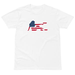 Könnte beinhalten: Weißes T-Shirt mit einer Grafik einer Person in Schussposition, die die Sterne und Streifen der amerikanischen Flagge verwendet. Das Design ist in Rot, Weiß und Blau gehalten. Ein lässiges T-Shirt.
