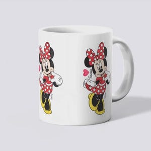 Puede incluir: Taza de cer&aacute;mica blanca con una ilustraci&oacute;n de dibujos animados de Minnie Mouse. Minnie est&aacute; representada con un vestido de lunares rojos, un lazo y zapatos. La taza tiene un asa curva y un dise&ntilde;o en ambos lados.