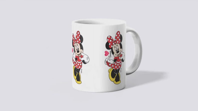Puede incluir: Taza de cer&aacute;mica blanca con una ilustraci&oacute;n de dibujos animados de Minnie Mouse. Minnie est&aacute; representada con un vestido de lunares rojos, un lazo y zapatos. La taza tiene un asa curva y un dise&ntilde;o en ambos lados.