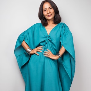 May include: A teal-colored caftan dress with wide sleeves and a tie detail at the neckline. The dress is floor-length and has a relaxed fit. The model is wearing silver bracelets and a watch.