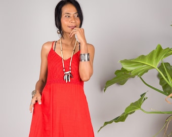 May include: A woman wearing a red maxi dress with a white beaded necklace and a silver bracelet. The dress has a simple design and is made of a lightweight fabric. The woman is standing in front of a gray wall with a plant in the background. The image includes the text "NUI CHAN © 2022 NUI CHAN".