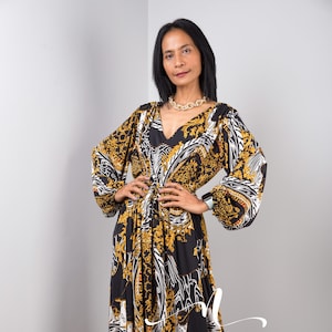 Puede incluir: Un vestido maxi negro con estampado barroco dorado y blanco. El vestido tiene mangas largas y un escote en V. El vestido está ajustado a la cintura con un cinturón de tela a juego.