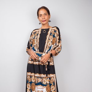 May include: A woman wearing a long black and blue maxi dress with a gold and white patterned design. The dress has a cinched waist and a flowing skirt.