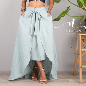May include: A woman wearing a blue halter top with a layered necklace and a light blue wrap skirt with a high-low hemline. The skirt has a tie at the waist. The woman is standing in front of a gray wall with a plant in the background.