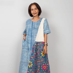 Op de afbeelding: Een knielange jurk met patchwork-ontwerp. De bovenste helft is blauw met een geometrisch patroon en een wit paneel aan de rechterkant. De onderste helft heeft een bloemenprint in blauw, roze en geel. De jurk heeft korte mouwen.