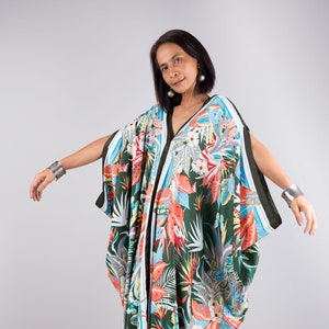 May include: A flowing, dark green caftan with a vibrant tropical floral print in shades of red, orange, blue, and white. The caftan has wide sleeves and a V-neckline. Black trim outlines the edges. The model is wearing silver bracelets.