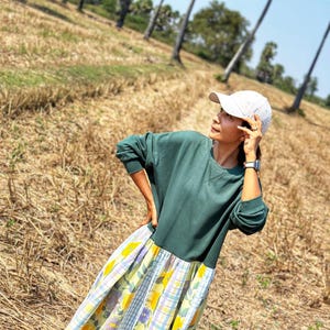 May include: A woman wearing a green long-sleeved shirt with a yellow and blue floral print skirt. She is wearing a white baseball cap and is standing in a field of dry grass.