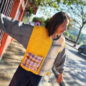Puede incluir: Una mujer lleva una blusa de patchwork multicolor con un panel amarillo, un panel azul y blanco y un panel morado y marrón. La blusa tiene un bolsillo grande en la parte delantera. La mujer lleva pantalones azul oscuro.