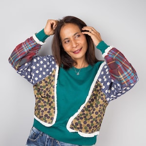 May include: A teal sweatshirt with patchwork sleeves. The sleeves feature a mix of patterns, including blue polka dots with white flowers, a camouflage-like design, and red plaid. The person is wearing blue jeans.