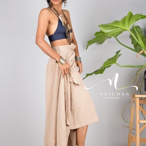 May include: A woman wearing a navy blue halter top and a beige wrap skirt. She is wearing a silver necklace and bracelets. The skirt is asymmetrical and has a high-low hemline. The woman is standing in front of a white wall with a green plant in the background.