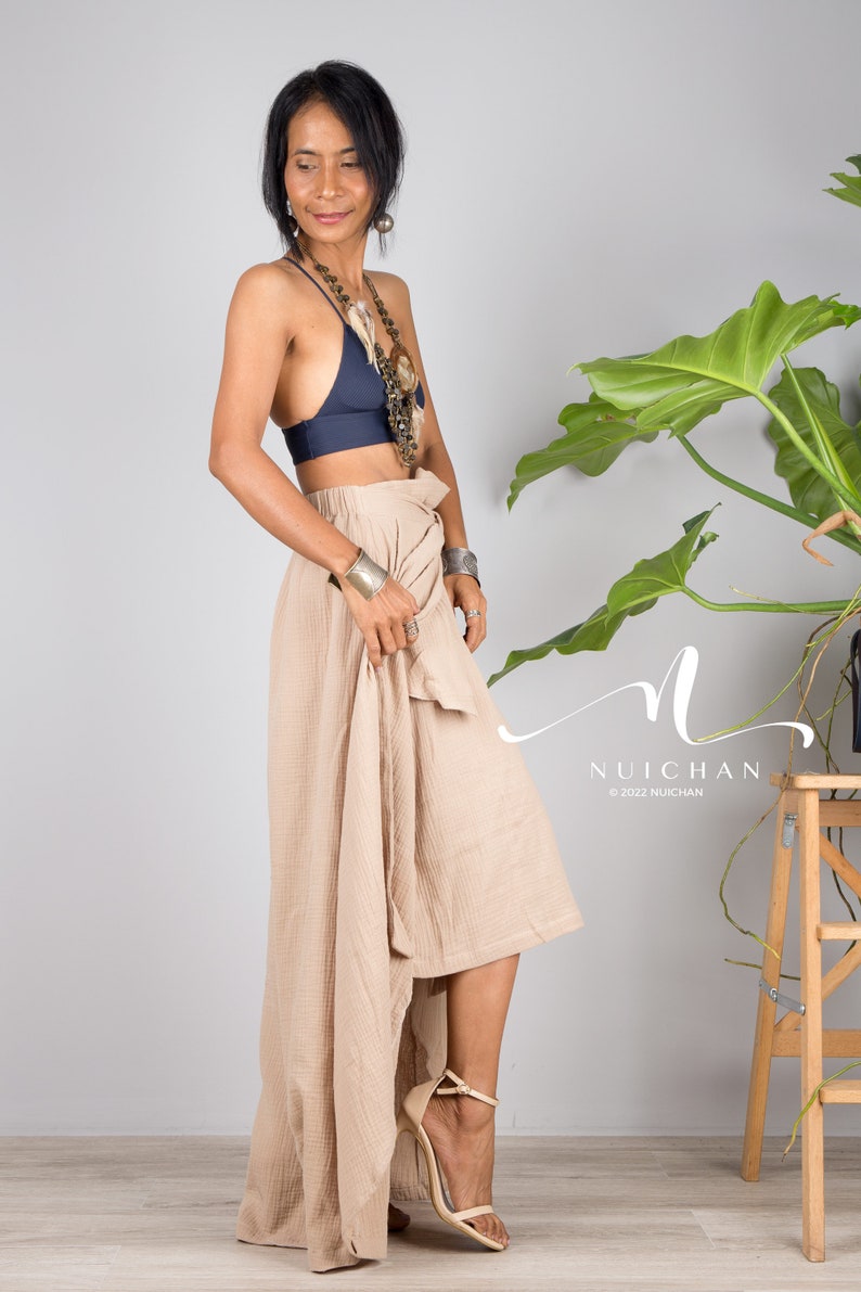 May include: A woman wearing a navy blue halter top and a beige wrap skirt. She is wearing a silver necklace and bracelets. The skirt is asymmetrical and has a high-low hemline. The woman is standing in front of a white wall with a green plant in the background.