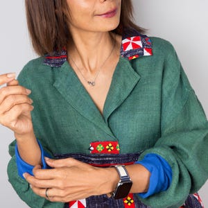 K&ouml;nnte beinhalten: Eine Frau tr&auml;gt eine gr&uuml;ne Leinenjacke mit bunten Stickereien. Die Jacke hat einen Kragen und lange &Auml;rmel. Die Frau tr&auml;gt eine silberne Halskette und eine Uhr am linken Handgelenk.