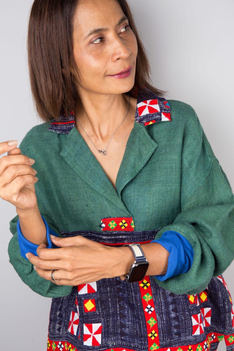 K&ouml;nnte beinhalten: Eine Frau tr&auml;gt eine gr&uuml;ne Leinenjacke mit bunten Stickereien. Die Jacke hat einen Kragen und lange &Auml;rmel. Die Frau tr&auml;gt eine silberne Halskette und eine Uhr am linken Handgelenk.