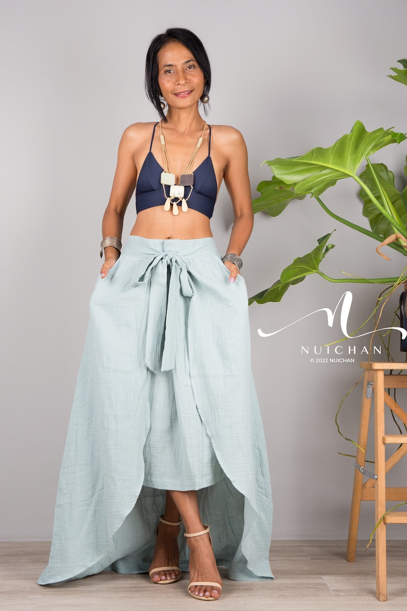 May include: A woman wearing a blue halter top with a layered necklace and a light blue wrap skirt with a high-low hemline. The skirt has a tie at the waist. The woman is standing in front of a gray wall with a plant in the background.