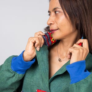 K&ouml;nnte beinhalten: Eine Frau tr&auml;gt eine gr&uuml;ne Jacke mit blauen und roten Verzierungen. Die Jacke hat ein farbenfrohes Stickmuster auf der Vorderseite. Sie tr&auml;gt eine silberne Halskette mit einem Herzanh&auml;nger.