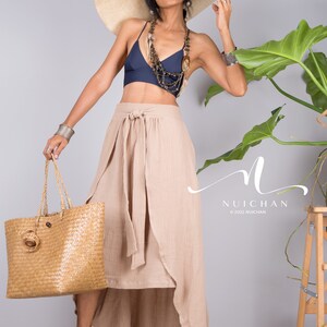 May include: A woman wearing a wide-brimmed straw hat, a navy blue halter top, a beige wrap skirt, and beige heeled sandals. She is holding a woven tote bag and standing in front of a grey wall with a potted plant.
