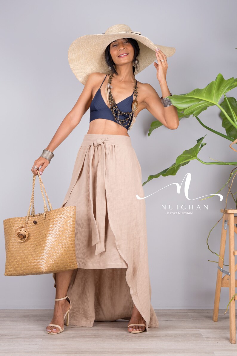 May include: A woman wearing a wide-brimmed straw hat, a navy blue halter top, a beige wrap skirt, and beige heeled sandals. She is holding a woven tote bag and standing in front of a grey wall with a potted plant.