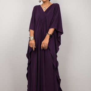 May include: A woman wearing a long, purple kaftan with a ruffled hem. She is wearing silver jewelry and sandals.