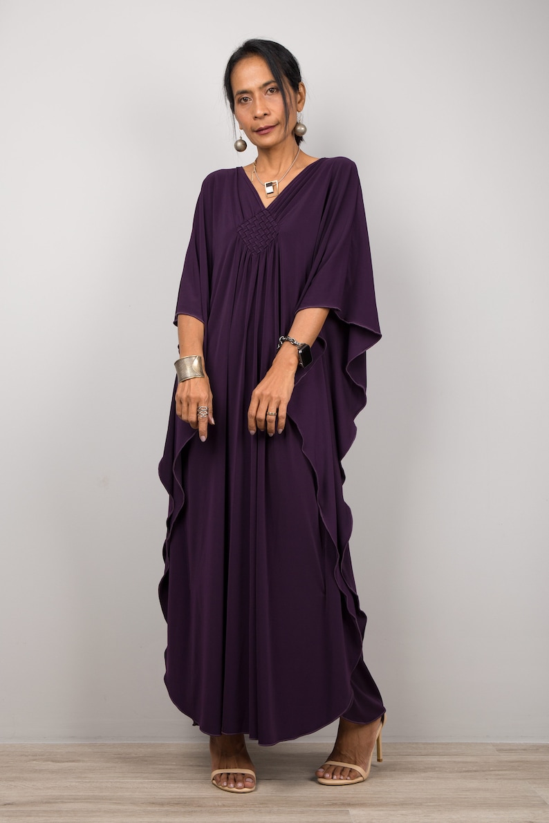 May include: A woman wearing a long, purple kaftan with a ruffled hem. She is wearing silver jewelry and sandals.