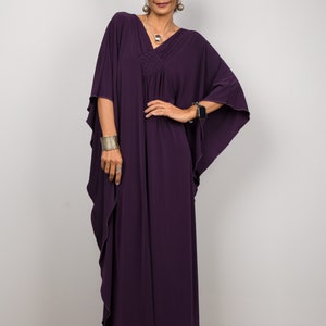 May include: A woman wearing a long, flowing purple kaftan with a V-neckline and a gathered waist. She is wearing silver earrings and a silver necklace.