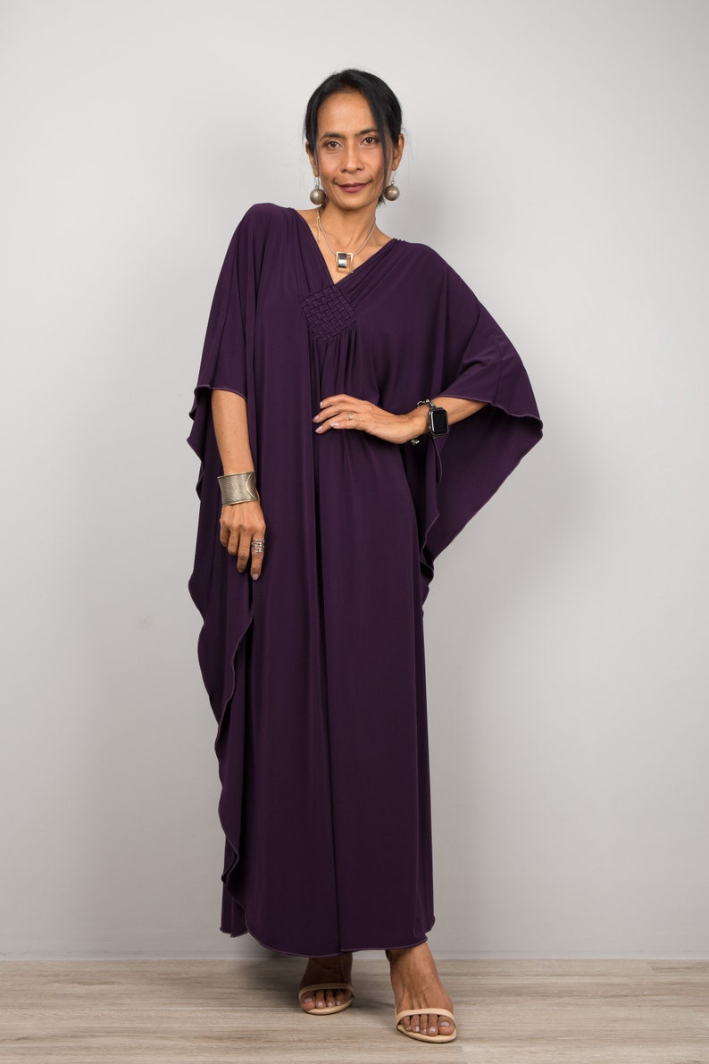 May include: A woman wearing a long, flowing purple kaftan with a V-neckline and a gathered waist. She is wearing silver earrings and a silver necklace.