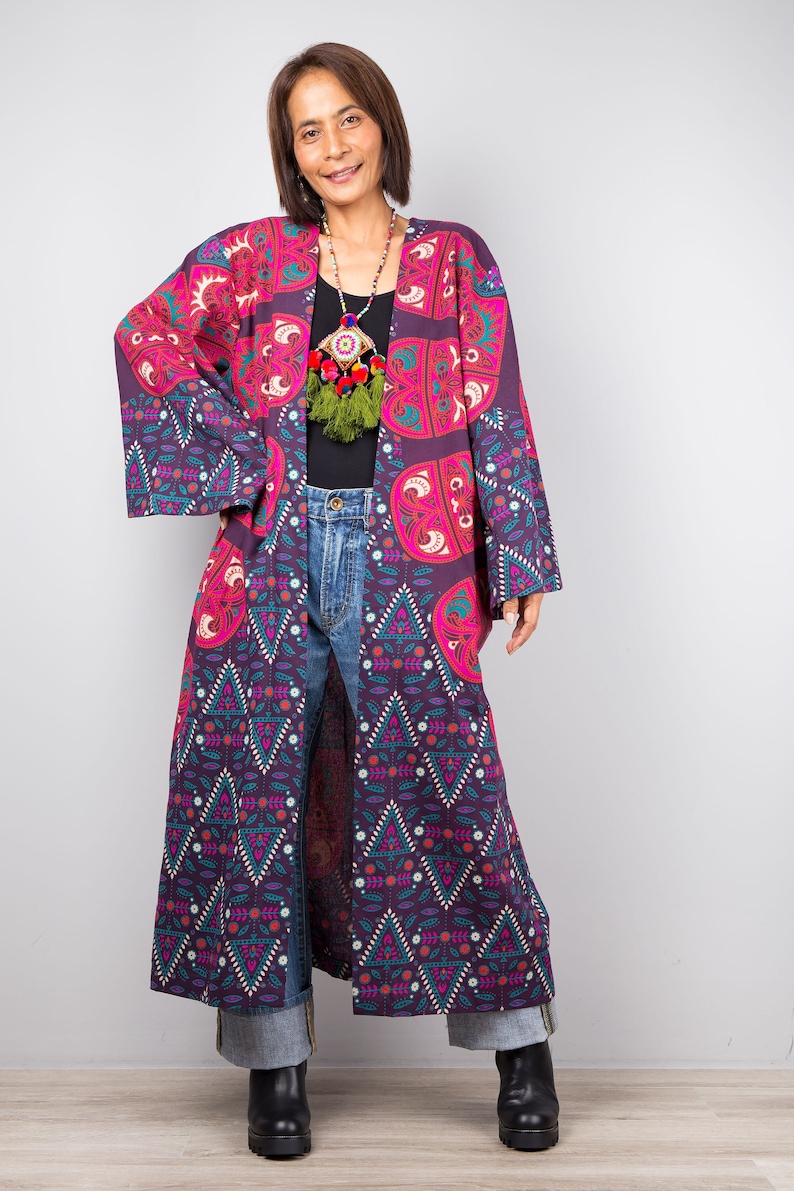 Pu&ograve; includere: Un kimono lungo con motivo viola e rosa con un design geometrico. Il kimono ha una manica larga e fluida e un orlo lungo e fluido. La modella indossa il kimono sopra una maglia nera e jeans blu.