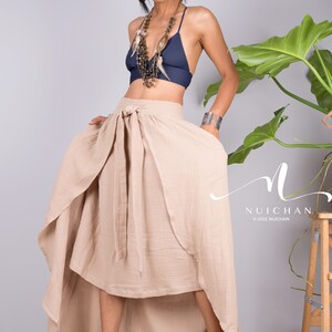 May include: A woman wearing a beige wrap skirt with a high-low hemline. The skirt is tied at the waist with a bow. She is also wearing a navy blue bra and a beaded necklace with feathers.
