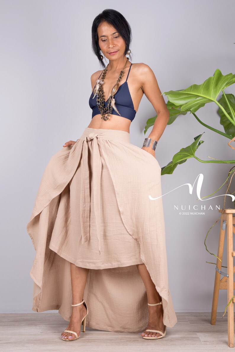 May include: A woman wearing a beige wrap skirt with a high-low hemline. The skirt is tied at the waist with a bow. She is also wearing a navy blue bra and a beaded necklace with feathers.