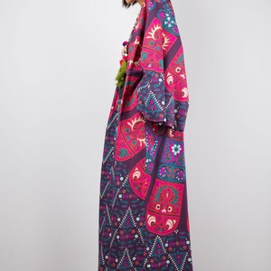 Pu&ograve; includere: Una lunga veste a maniche lunghe con un motivo geometrico a fiori in viola e rosa. La veste ha un collo ampio e aperto. La modella indossa la veste con stivali neri.