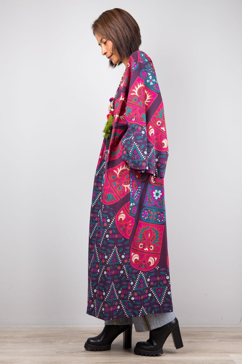 Pu&ograve; includere: Una lunga veste a maniche lunghe con un motivo geometrico a fiori in viola e rosa. La veste ha un collo ampio e aperto. La modella indossa la veste con stivali neri.