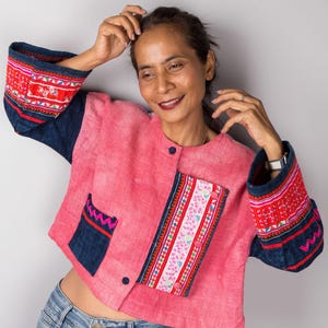 Könnte beinhalten: Eine Frau trägt eine rosafarbene, kurze Jacke mit blauen und roten Stickereien. Die Jacke hat einen Knopfverschluss und eine Tasche auf der linken Seite. Sie trägt auch eine blaue Jeans.