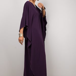 May include: A woman wearing a long, flowing purple kaftan with a ruffled hem. She is wearing silver jewelry and has her hair pulled back.