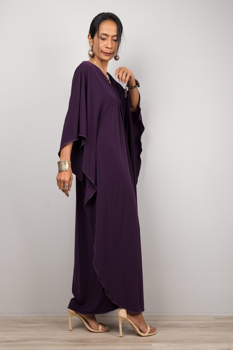 May include: A woman wearing a long, flowing purple kaftan with a ruffled hem. She is wearing silver jewelry and has her hair pulled back.