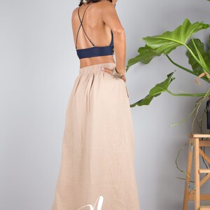 May include: A woman wearing a navy blue crop top with a criss-cross back and a beige maxi skirt. The skirt has a gathered waist and a flowing silhouette. The woman is standing in front of a white wall with a potted plant in the background.