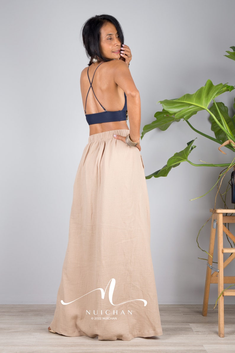 May include: A woman wearing a navy blue crop top with a criss-cross back and a beige maxi skirt. The skirt has a gathered waist and a flowing silhouette. The woman is standing in front of a white wall with a potted plant in the background.