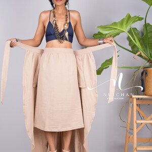 May include: A woman wearing a beige wrap skirt with a long train, a navy blue bra, and a layered necklace. The skirt is tied at the waist and has a high-low hemline. The woman is standing in front of a grey background with a potted plant to the right.